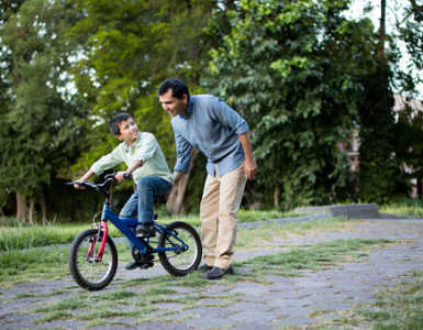 Ein Junge lernt Fahrradfahren, sein Vater schiebt ihn mit einer Hand an. Im Hintergrund sind Bäume zu sehen.