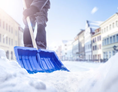 Eine Person benutzt eine Schneeschaufel auf einer verschneiten Straße. Im Hintergrund sieht man einige Häuser.