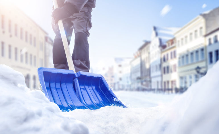 Eine Person benutzt eine Schneeschaufel auf einer verschneiten Straße. Im Hintergrund sieht man einige Häuser.