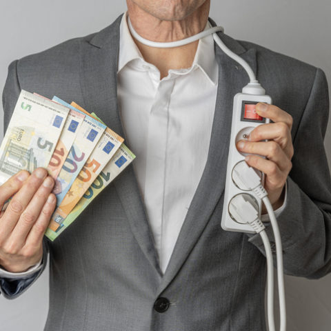 Ein Mann im Anzug hält in einer Hand mehrere Euro-Scheine, in der anderen eine Mehrfachsteckdose. Das Kabel ist um seinen Hals gewickelt.