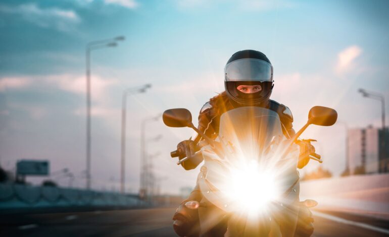 Ein Motorradfahrer mit Schutzkleidung fährt auf der Straße auf die Kamera zu, das Licht blendet. Dahinter hohe Straßenlaternen.
