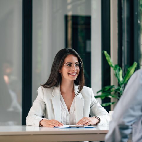 Eine Geschäftsfrau mit weißem Blazer und Brille stellt einem Mann in Hemd und Brille Fragen im Bewerbungsgespräch. Zwischen ihnen ist ein Tisch mit Unterlagen, dahinter eine Glasfront.