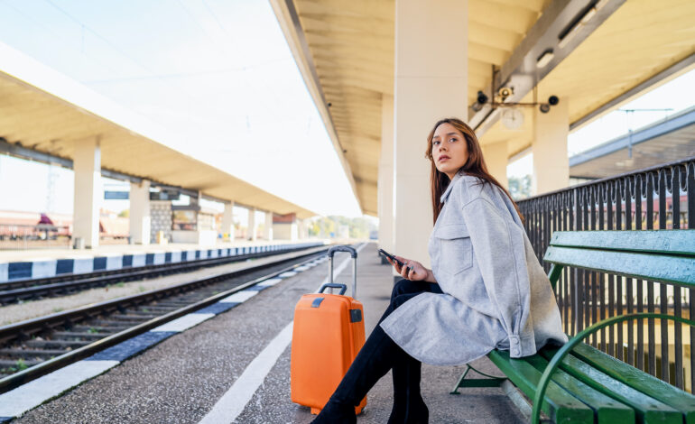 Frau sitzt am Bahnsteig, prüft mit dem Smartphone ihre Rechte bei Zugverspätung, neben sich einen orangefarbenen Koffer.