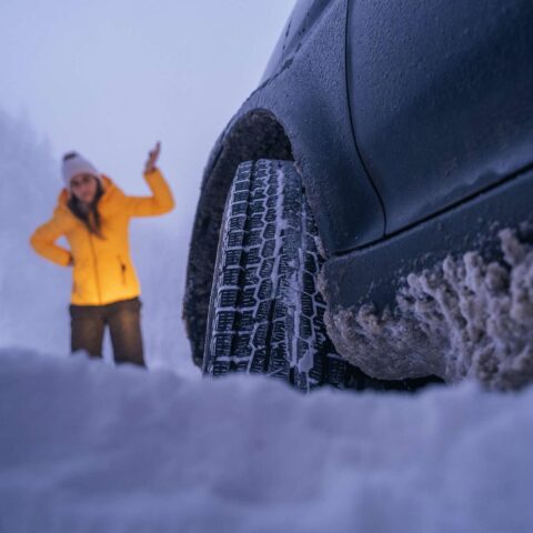 Frau mit Mütze und Winterjacke neben festgefahrenem Auto im Winter.