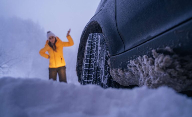 Frau mit Mütze und Winterjacke neben festgefahrenem Auto im Winter.