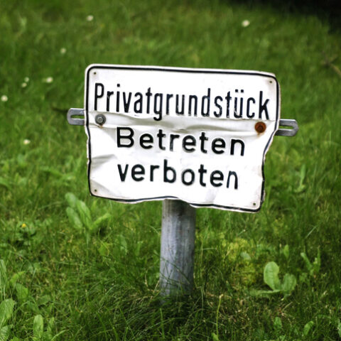 Ein kleines verbeultes Schild auf einer Wiese, das Hausfriedensbruch vorbeugen soll. Darauf steht Privatgrundstück, Betreten verboten.
