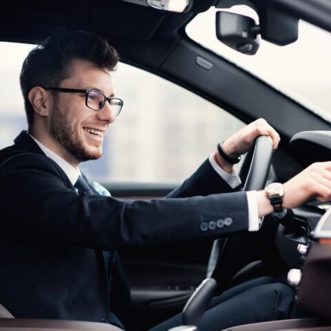 Mann mit Brille und Anzug im Auto mit einer Hand am Leckrad und einer Hand am Touchscreen während der Fahrt.