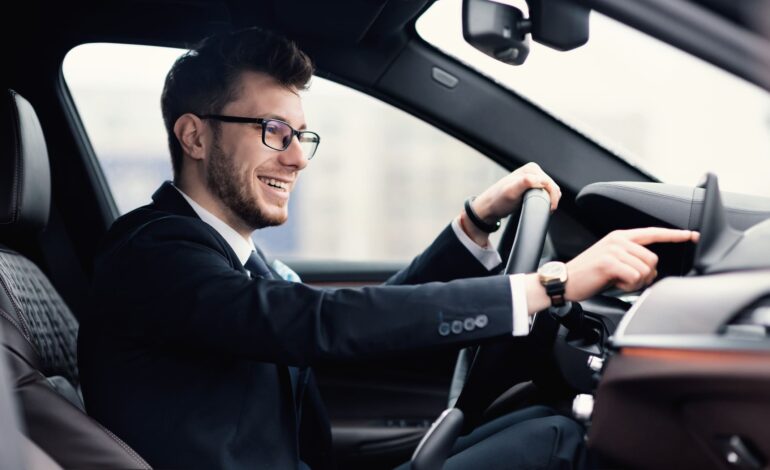 Mann mit Brille und Anzug im Auto mit einer Hand am Leckrad und einer Hand am Touchscreen während der Fahrt.