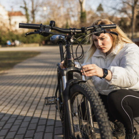 Frau mit weißer Jacke hockt auf einem gepflasterten Weg und macht einen Fahrrad Frühjahrscheck, sie überprüft etwas am Vorderreifen.
