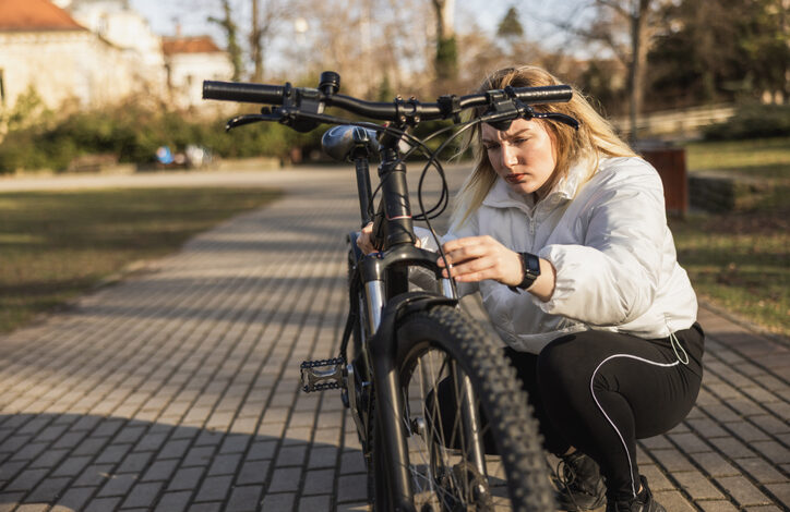 Frau mit weißer Jacke hockt auf einem gepflasterten Weg und macht einen Fahrrad Frühjahrscheck, sie überprüft etwas am Vorderreifen.