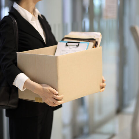 Frau mit Bluse und Blazer hält eine Kiste mit Büromaterial in den Händen, nach der Kündigung durch den Arbeitgeber