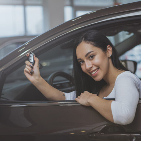 Junge Frau mit bestandenem Führerschein sitzt im Auto und zeigt ihren Autoschlüssel durch das geöffnete Fenster.
