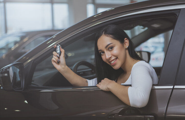 Junge Frau mit bestandenem Führerschein sitzt im Auto und zeigt ihren Autoschlüssel durch das geöffnete Fenster.
