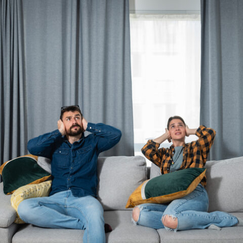 Ein Mann und eine Frau an Sonn- und Feiertagen auf einem Sofa vor einem Fenster mit Vorhängen. Beide halten sich die Ohren zu und blicken nach oben, wegen der Lärmbelästigung.