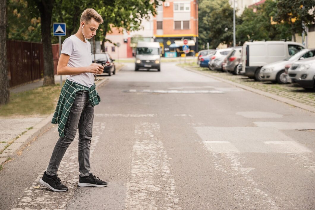Junger Mann überquert die Straße und schaut auf sein Smartphone. Dahinter nähert sich ein großes Fahrzeug. So geschieht ein Unfall mit Fußgänger.