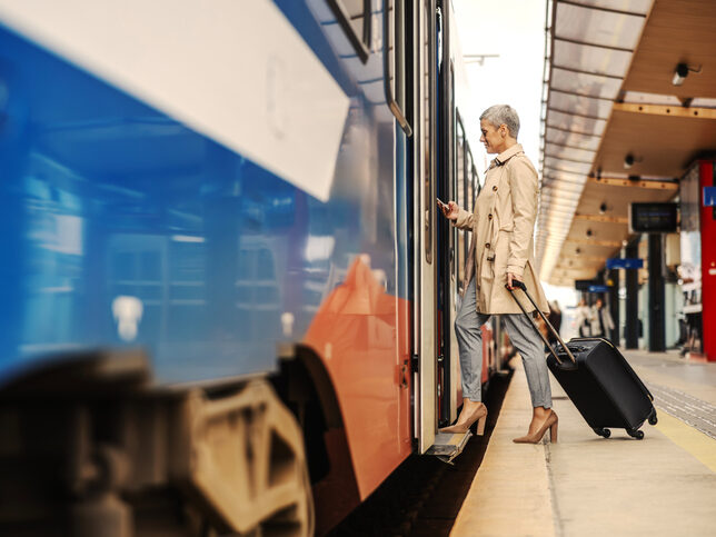 Eine ältere Frau mit einem Koffer, die in einen Zug steigt und dabei mit dem Smartphone den Fahrplan mit den Neuerungen ab Dezember 2025 checkt.