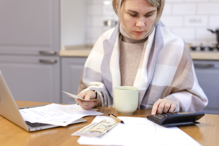 Frau sitzt am Küchentisch und dokumentiert die Mietminderung bei Heizungsausfall. Auf dem Tisch sind ein Laptop, ein Taschenrechner, eine Tasse, Dokumente, ein Stift und Geldscheine.