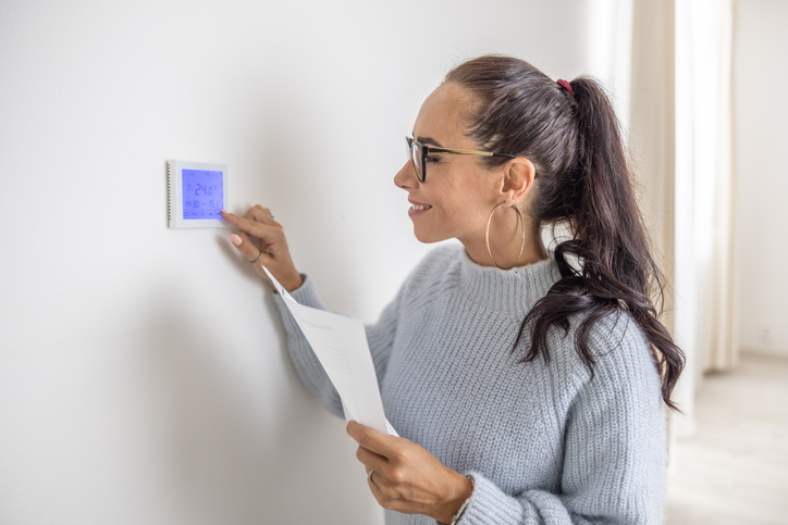 Lächelnde Mieterin bedient den Thermostat an der Wand, hält dabei ein Blatt Papier in der Hand.