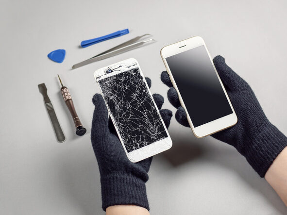 Zwei behandschuhte Hände, in jeder Hand ein Smartphone. Links ein defektes Display, rechts ein intaktes. Im Hintergrund Werkzeuge zur Reparatur.