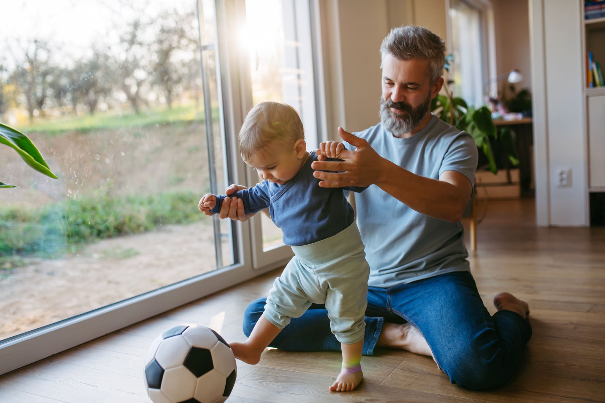 Mann während der Elternzeit, hält sein Kleinkind an den Händen, das gegen einen Fußball tritt.