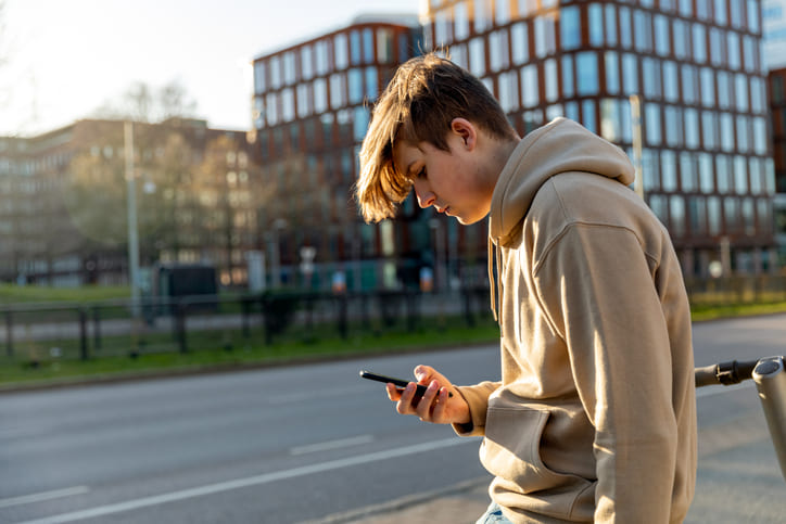 Junge im Alter von 14 mit Hoodie und Blick auf sein Smartphone. Im Hintergrund eine leere Straße und Hochhäuser.