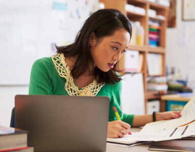 Eine Frau mit Stift und Papieren in den Händen sitzt an einem Schreibtisch beim Arbeitszeugnis prüfen. Daneben steht ein Laptop.