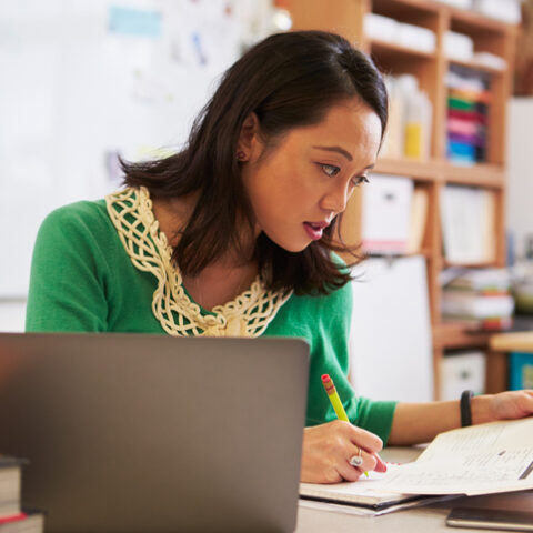 Eine Frau mit Stift und Papieren in den Händen sitzt an einem Schreibtisch beim Arbeitszeugnis prüfen. Daneben steht ein Laptop.