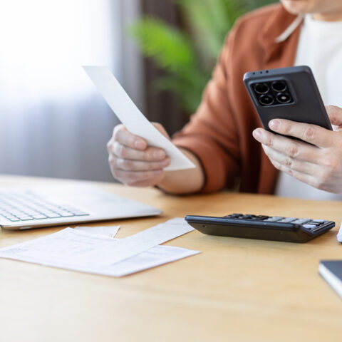Jemand sitzt an einem Schreibtisch, in einer Hand eine Rechnung, in der anderen Hand ein Smartphone. Auf dem Tisch liegen potenzielle Fake Rechnungen und ein Laptop.