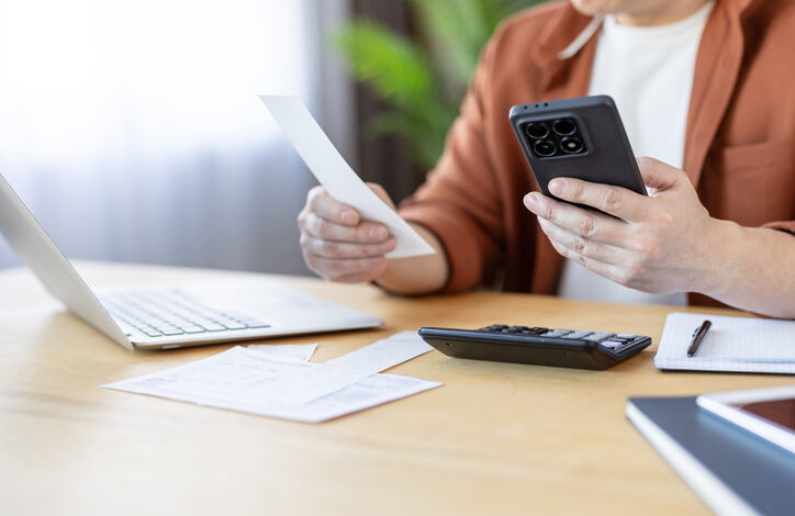 Jemand sitzt an einem Schreibtisch, in einer Hand eine Rechnung, in der anderen Hand ein Smartphone. Auf dem Tisch liegen potenzielle Fake Rechnungen und ein Laptop.