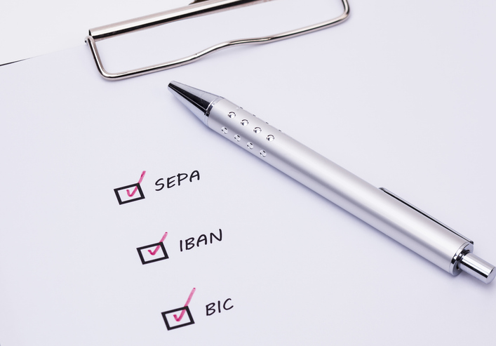 Ein silberner Kugelschreiber liegt auf einem Papier auf einem Klemmbrett. Dort sind abgehakte Kästchen, daneben steht SEPA, IBAN und BIC.