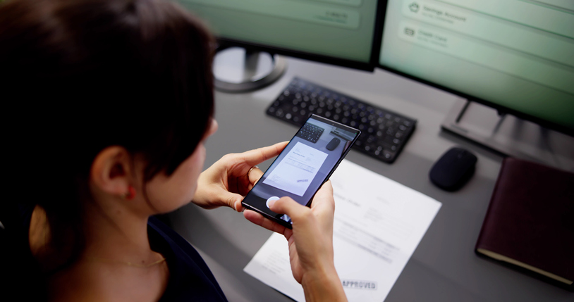 Eine Frau von hinten, die an einem Schreibtisch vor zwei Bildschirmen sitzt. Sie scannt mit dem Smartphone potenzielle Fake Rechnungen.