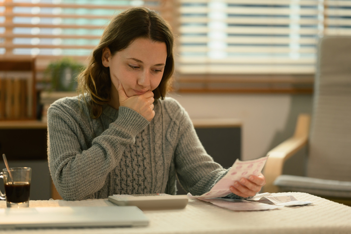Junge Frau in Pullover hält nachdenklich die Hand am Kinn und prüft an ihrem Schreibtisch potenzielle Fake Rechnungen. Daneben steht ein Glas mit Kaffee, dahinter sind Jalousien zu sehen.