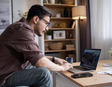 Mann mit Brille im Homeoffice sitzt auf der Couch am Couchtisch mit einem Laptop, einem Smartphone und einer Tasse. Dahinter ein Regal und eine Stehlampe