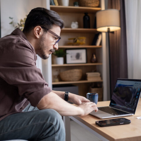 Mann mit Brille im Homeoffice sitzt auf der Couch am Couchtisch mit einem Laptop, einem Smartphone und einer Tasse. Dahinter ein Regal und eine Stehlampe