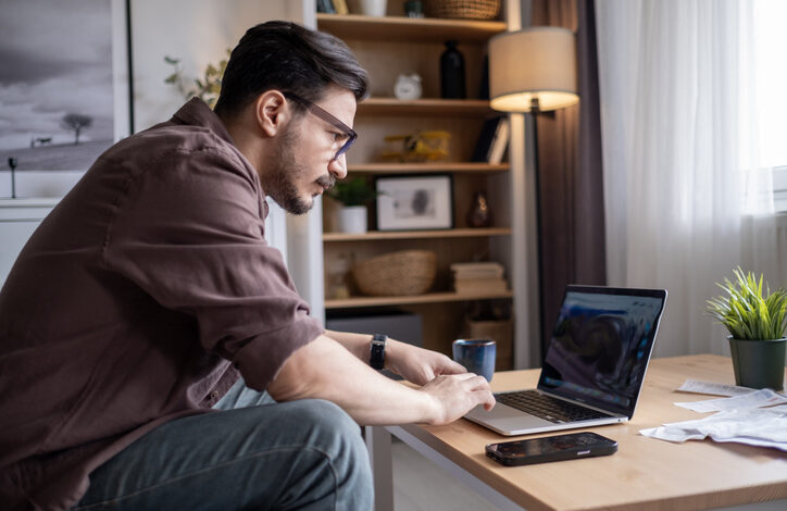 Mann mit Brille im Homeoffice sitzt auf der Couch am Couchtisch mit einem Laptop, einem Smartphone und einer Tasse. Dahinter ein Regal und eine Stehlampe