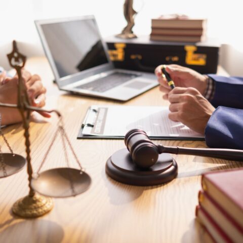 Ansicht von Armen und Händen zweier Männer, die sich an einem Tisch gegenüber sitzen. Der eine ist Anwalt, der andere möchte den passenden Anwalt finden. Dazwischen ein Richterhammer, eine Goldwaage, ein Laptop, Bücher und Dokumente.