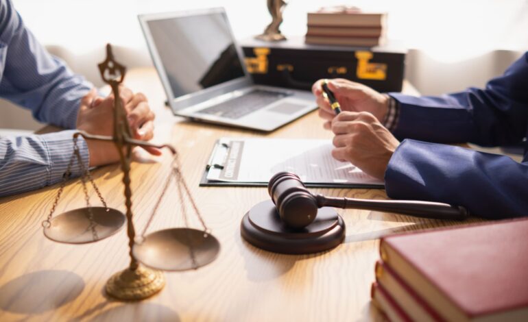 Ansicht von Armen und Händen zweier Männer, die sich an einem Tisch gegenüber sitzen. Der eine ist Anwalt, der andere möchte den passenden Anwalt finden. Dazwischen ein Richterhammer, eine Goldwaage, ein Laptop, Bücher und Dokumente.