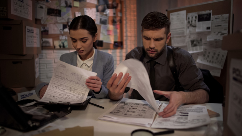 Eine Frau und ein Mann wollen einen passenden Anwalt finden, sie sitzen an einem Schreibtisch und blättern Papierstapel durch.