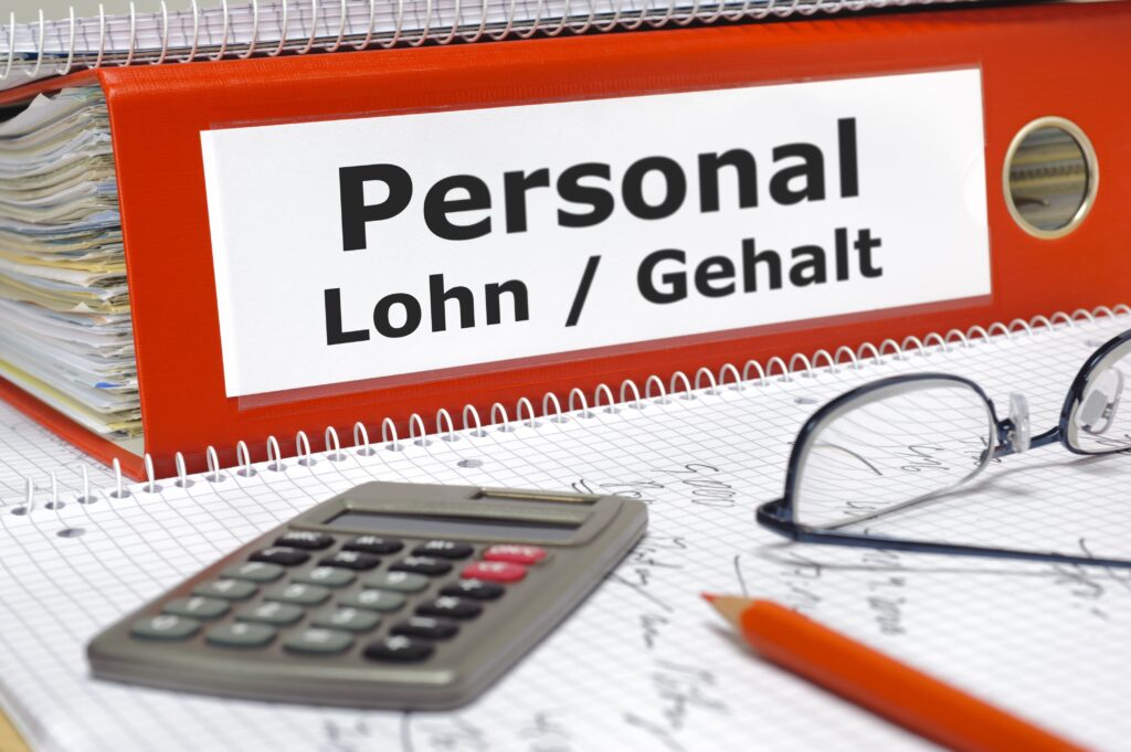 Ein roter Ordner mit den Neuerungen Juni 2026 in Personal Lohn / Gehalt, davor Papier, Stift, Taschenrechner und Brille.