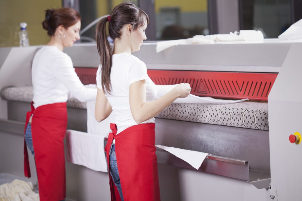 Was darf man mit 16? Zwei Junge Frauen mit zusammengebundenen Haaren, weißen Shirts und roten Schürzen bei der Arbeit an einer großen Bügelmaschine.