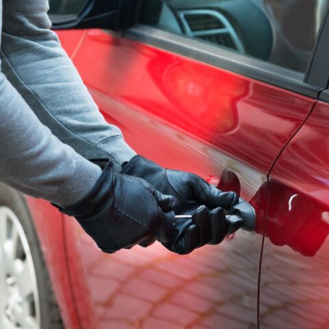 Hier wird ein rotes Auto gestohlen von einem Mann, der mit behandschuhten Händen die Tür aufbrechen will.