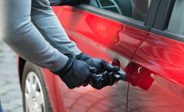 Hier wird ein rotes Auto gestohlen von einem Mann, der mit behandschuhten Händen die Tür aufbrechen will.