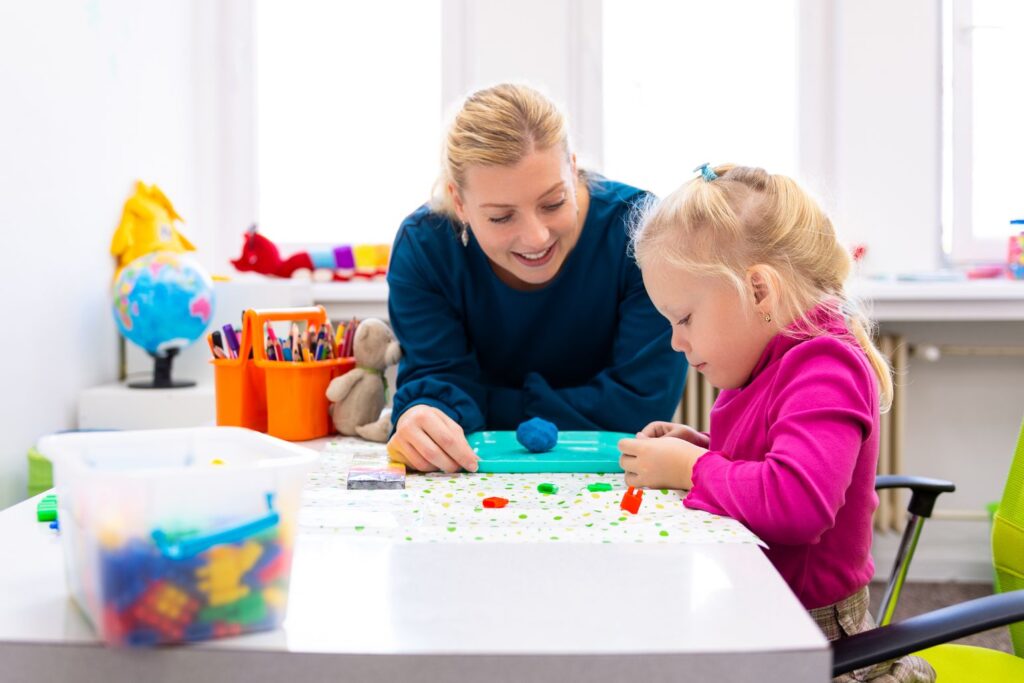 Eine blonde Frau bei der Betreuung eines blonden Mädchens. Auch hier gibt es Neuerungen im August 2026. Die beiden kneten an einem kleinen Tisch.