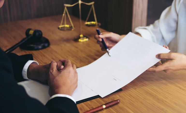 Die Hände zweier Personen am Tisch. Eine Person hält der anderen ein Papier und einen Stift zum Unterschreiben der Erstattung der außergerichtlichen Anwaltskosten hin. Dahinter ein Richterhammer und eine goldene Waage.