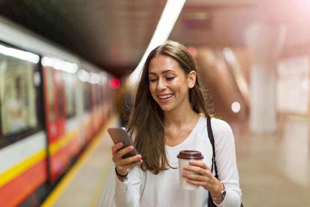 Eine Frau am Bahnhof, die auf ihrem Handy die Neuerungen Oktober 2026 zum Alkoholverbot checkt. In der anderen Hand hält sie einen ToGo-Becher, hinter ihr ist eine U-Bahn zu sehen.