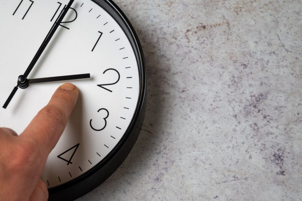 Eine große Uhr auf grauem Hintergrund und ein Finger, der darauf zeigt.