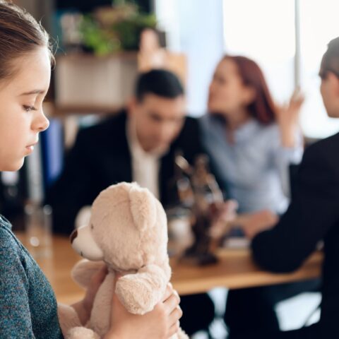 Mädchen mit Teddybär im Vordergrund. Dahinter Mutter und Vater, die mit Anwalt über Unterhalt diskutieren.