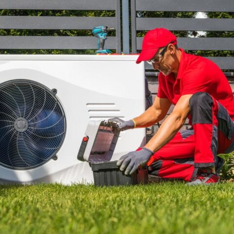 Nach § 14a EnWG baut ein Netzbetreiber in roter Arbeitskleidung ein Außengerät einer Wärmepumpe um. Er hat einen Werkzeugkoffer dabei und auf dem Gerät ist ein Akkuschrauber abgestellt.